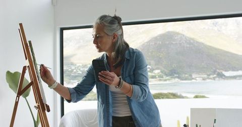 Mature woman painting on easel holding smartphone with coastal view through large window