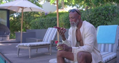 Senior man relaxing poolside holding cocktail and scrolling smartphone in linen shirt