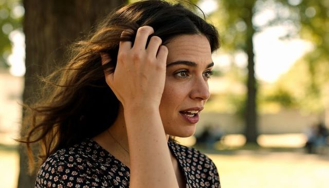 Young woman touching hair and speaking in sunlit park wearing floral blouse and necklace