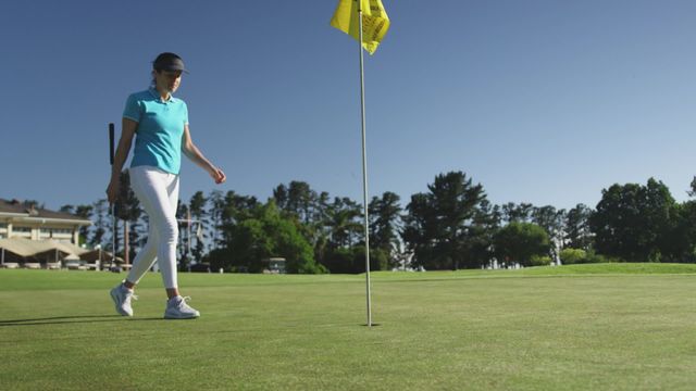 Female golfer on lush, green golf course retrieving a golf ball from the hole. She wears a blue polo shirt, white pants, and a cap. Trees and a clubhouse are visible in the background against a clear blue sky. Ideal for promoting outdoor sports activities, healthy lifestyles, athletic wear, golfing equipment, and leisure activities.