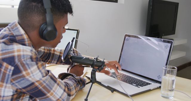 Man wearing headphones recording a podcast using a laptop and a microphone. Ideal for illustrating home office environments, podcast creation, remote work setups, and digital content production.