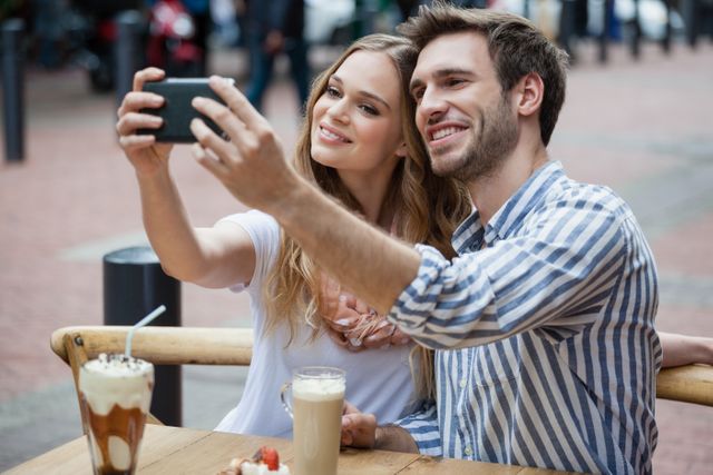 Happy couple holding smart phone while sitting at sidewalk cafe in city