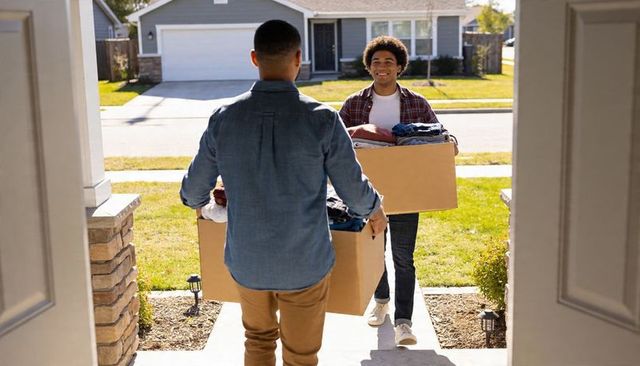 Carrying moving boxes onto suburban front porch while smiling, friends helping with move