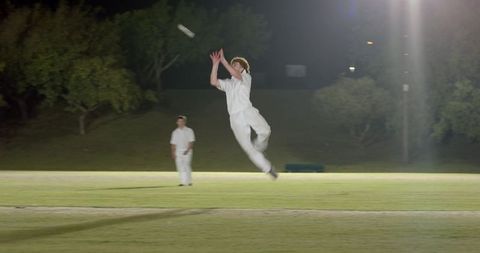 Leaping cricketer catching white ball under floodlights at night on grass pitch