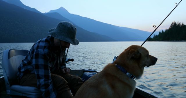 Man fishing in boat with dog, mountains silhouetted in background, lake reflecting evening light. Ideal for themes related to nature, leisure, outdoor activities, peaceful retreats, and human-animal bonds.