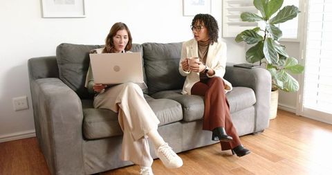 Diverse women collaborating on laptop while drinking coffee in bright home office lounge