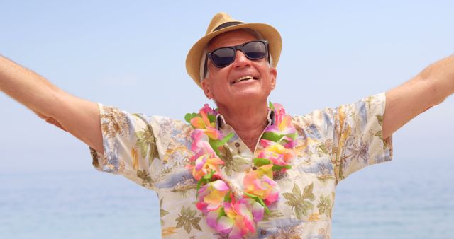 This stock photo depicts a smiling senior man wearing a Hawaiian shirt, lei, hat, and sunglasses, standing with outstretched arms at a beach, conveying a sense of happiness and relaxation. Ideal for travel agencies, retirement advertisements, tropical vacation promotions, and lifestyle blogs focusing on senior adventures and happiness.