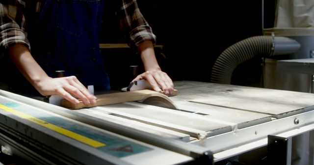 Hands of a woodworker skillfully operating a table saw in a workshop. Ideal for use in articles, blogs, or advertisements focusing on craftsmanship, woodworking tutorials, DIY home projects, or promoting carpentry tools and safety equipment.