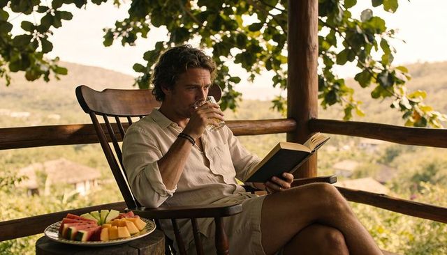 Man in linen shirt reading on rustic veranda rocking chair while sipping drink