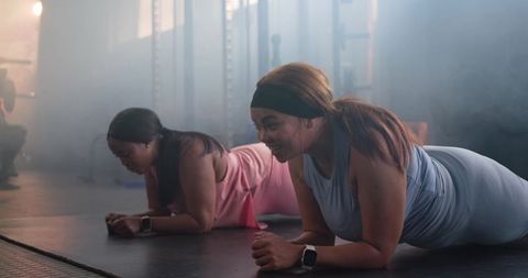 Diverse women performing forearm planks with smartwatches and battle rope in gym