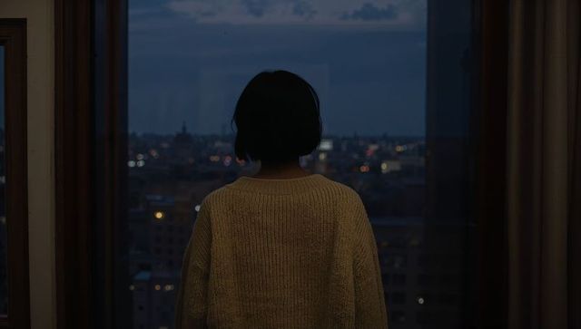 Woman gazing through floor-to-ceiling window at dusk wearing chunky knit sweater