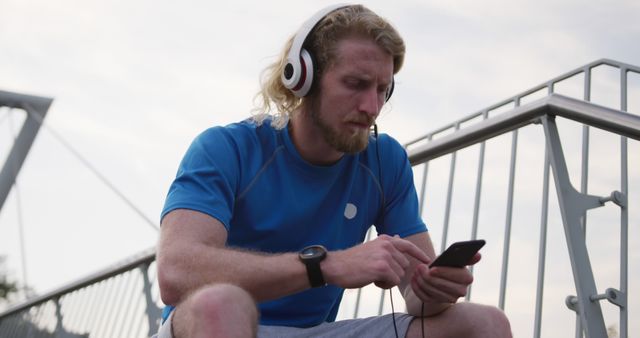 Young man sitting outdoors with headphones and listening to music on smartphone. Ideal for use in content related to lifestyle, relaxation, technology, audio entertainment, and urban culture.