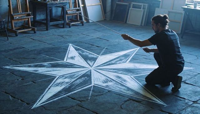Kneeling artist pulling string and drawing large chalk star on cracked studio floor
