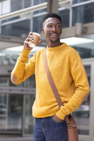 Smiling african commuter holding takeaway coffee wearing mustard sweater and crossbody bag