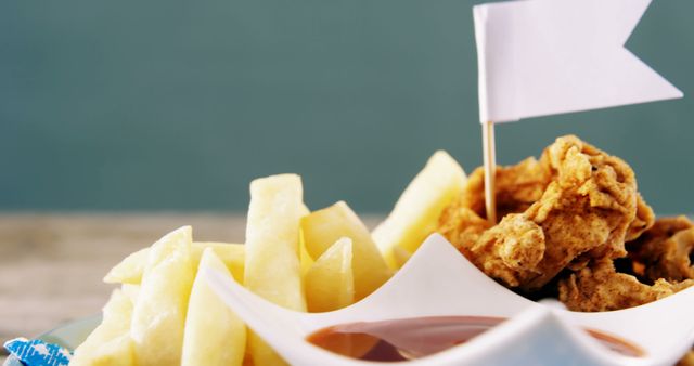 A serving of crispy fried chicken and golden french fries is presented in a paper container, with a white flag for added decoration. The combination of these popular fast-food items makes for a tempting meal option.