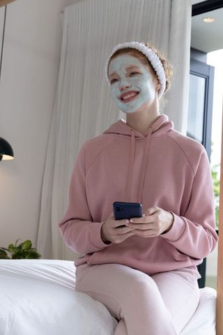 Young woman wearing mint face mask and pink loungewear sitting on bed holding smartphone