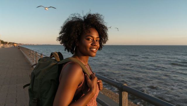 Smiling young woman on coastal boardwalk at golden hour carrying olive green backpack