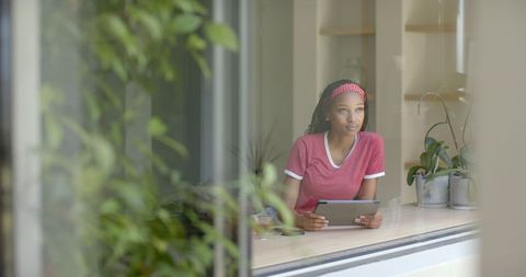 Thoughtful young woman by window holding tablet