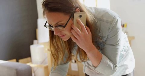 Young woman leaning over packing boxes while talking on smartphone in sunlit living room