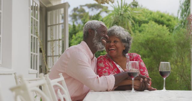 Senior biracial couple enjoys a romantic moment outdoors, with copy space. They share laughter over wine in a serene garden setting.