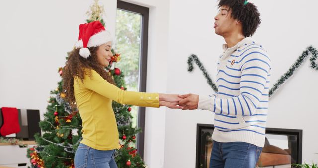 Image of happy diverse couple in reindeer antlers and santa hat dancing by christmas tree at home. Christmas, celebration, happiness, tradition and inclusivity concept.