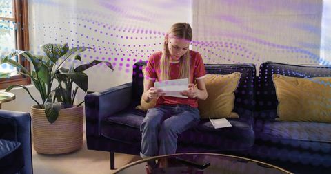 Young woman reading personal letter while sitting on dark sofa in cozy modern living room