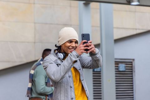 Young man smiling taking smartphone selfie wearing beanie and headphones on urban walkway