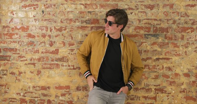 Caucasian man poses confidently against a brick wall. His casual style and sunglasses give off a relaxed, urban vibe.