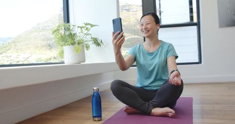 Asian woman taking yoga selfie while meditating on purple mat beside sunlit window