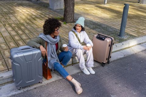 Multiracial friends sharing coffee and conversation while waiting with suitcases on city curb