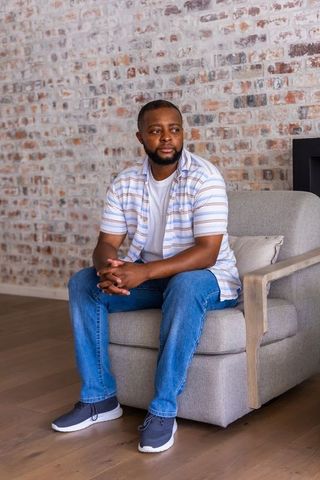 African american man sitting on armchair in minimalist living room with exposed brick