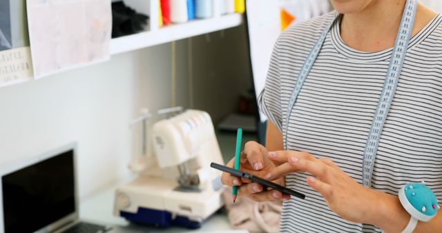 Image of tailor using smartphone in sewing workshop. Person is surrounded by sewing tools including a sewing machine and measuring tape. Ideal for use in articles about modern sewing techniques, small business management, or integrating technology in traditional crafts.