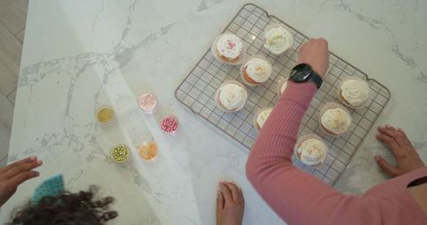Overhead family decorating buttercream cupcakes on marble countertop with pastel sprinkles