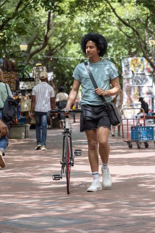 African american man walking pushing bicycle in tree-lined market wearing messenger bag