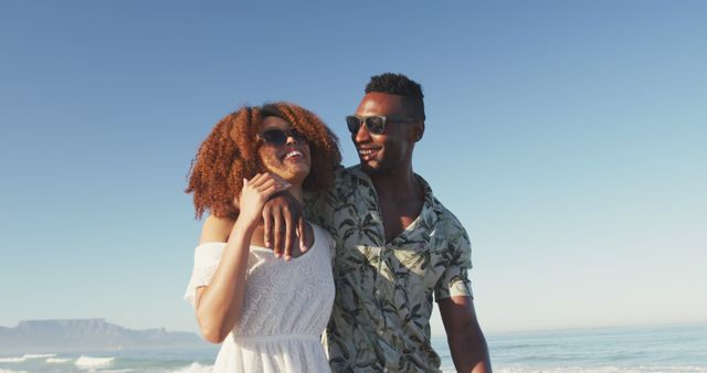 Happy diverse couple in sunglasses embracing and walking on sunny beach by the sea. Summer, free time, relaxation, romance and vacations.