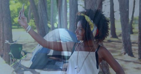 Smiling woman taking selfie at sunlit pine campsite with tent and summer accessories
