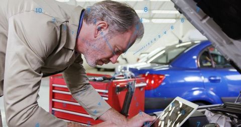 Senior mechanic leaning over engine inspecting with tablet in modern auto repair shop bay