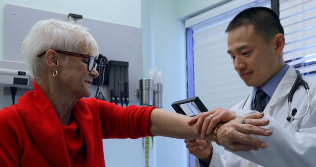 Senior woman sitting in medical office while male doctor with stethoscope examining her arm using medical device. Ideal for use in healthcare articles, medical publications, and brochures about medical check-ups, diagnostic procedures, or patient care.