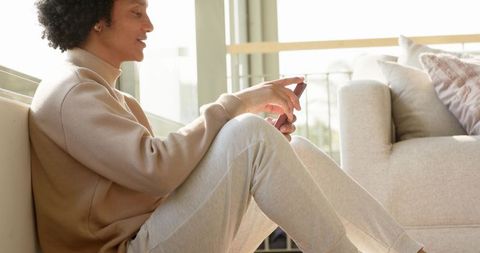 African american man relaxing on floor holding smartphone in minimalist bright living room