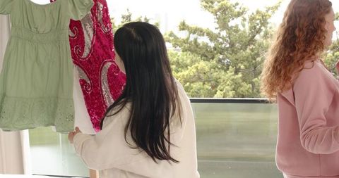 Diverse friends holding dresses on balcony browsing casual green dress and sequin gown