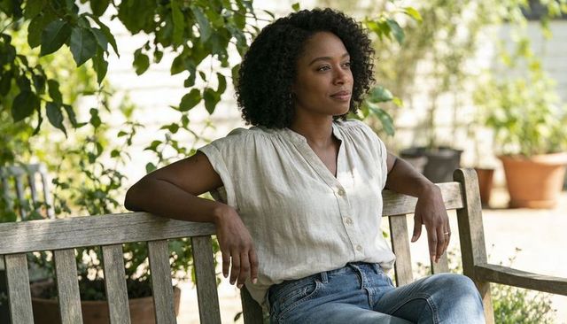 Relaxed woman sitting on wooden bench in shaded garden wearing linen blouse and jeans