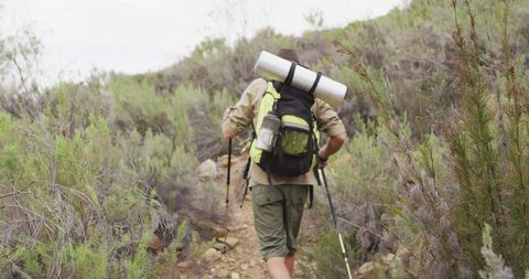 Solo hiker trekking uphill on rocky scrubland trail with loaded backpack and poles