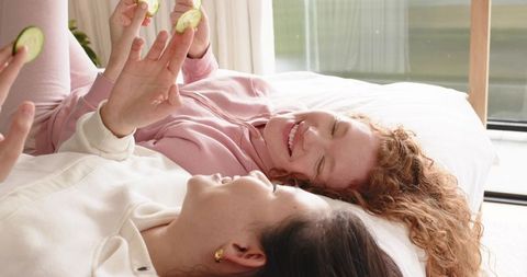 Cozy at-home spa moment with diverse friends lying on bed holding cucumber slices in soft loungewear