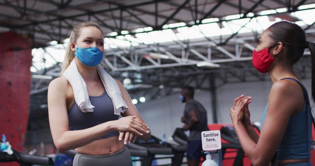 Two active women are wearing face masks in a gym while socializing. Each has a towel, and they are near treadmills with a hand sanitizer station in the foreground. This image can be used for promoting health safety during fitness activities, social interactions in gyms, or campaigns about exercising. Suitable for fitness centers, social distancing practices, and wellness campaigns.