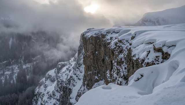 Snowy cliff rim revealing layered rock strata and wind-sculpted snowdrifts with hikers