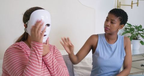 African american woman adjusting led facial mask on friend during cozy bedside skincare session