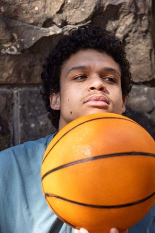 Sunlit urban portrait of young man leaning against stone wall holding basketball reflecting