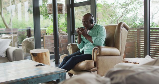 Middle-aged man seated on armchair, wearing headphones and drinking coffee. Room features warm, natural lighting, rustic wooden furniture, large windows with a view of greenery. Ideal for themes of relaxation, comfort, leisure, home lifestyle, elderly relaxation.