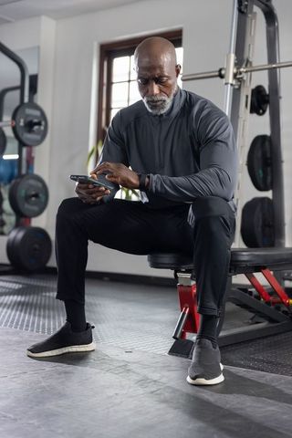 Senior man checking fitness tracker on smartphone in gym, sitting on bench tracking progress