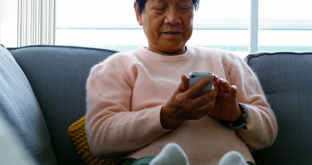 An elderly woman is casually sitting on a gray couch, using her smartphone in a bright living room. This image is useful for illustrating themes related to senior citizens embracing technology, staying connected through digital means, and enjoying comfortable, relaxed moments at home. It can be used in articles, blog posts, or advertisements promoting the use of technology among older adults, especially in the context of communication and the importance of digital literacy for seniors.